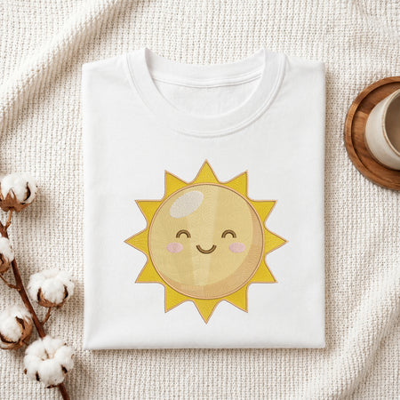 A folded white T-shirt with a cute embroidered-style smiling sun graphic in yellow, beige, and pink, styled on a textured cream fabric with cotton bolls and a small cup in the corners.