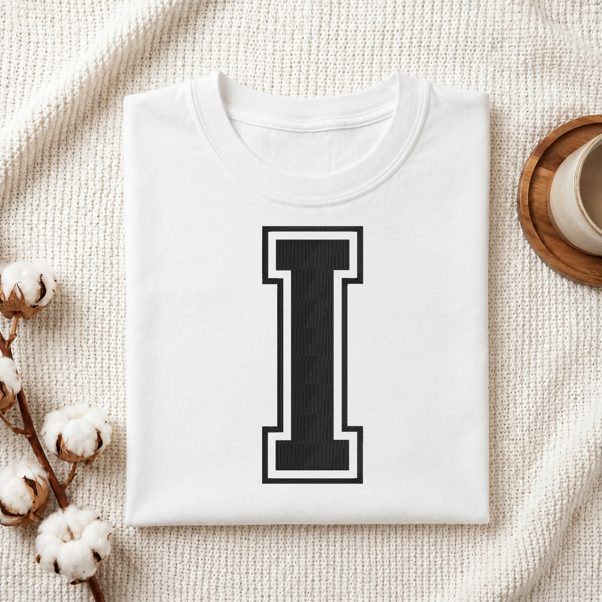 A folded white T-shirt with a large black block letter 'I' outlined in white on the front, styled on a textured cream fabric with cotton stems and a cup in the background.