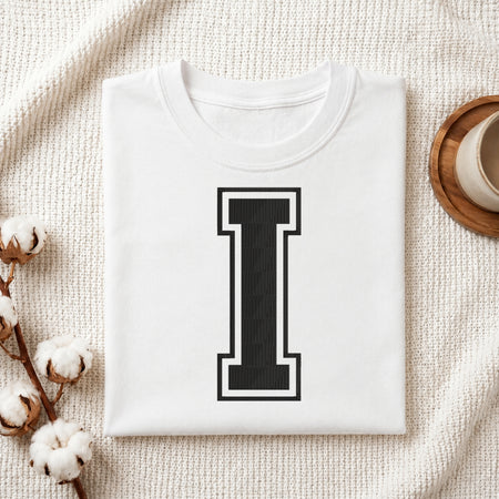 A folded white T-shirt with a large black block letter 'I' outlined in white on the front, styled on a textured cream fabric with cotton stems and a cup in the background.