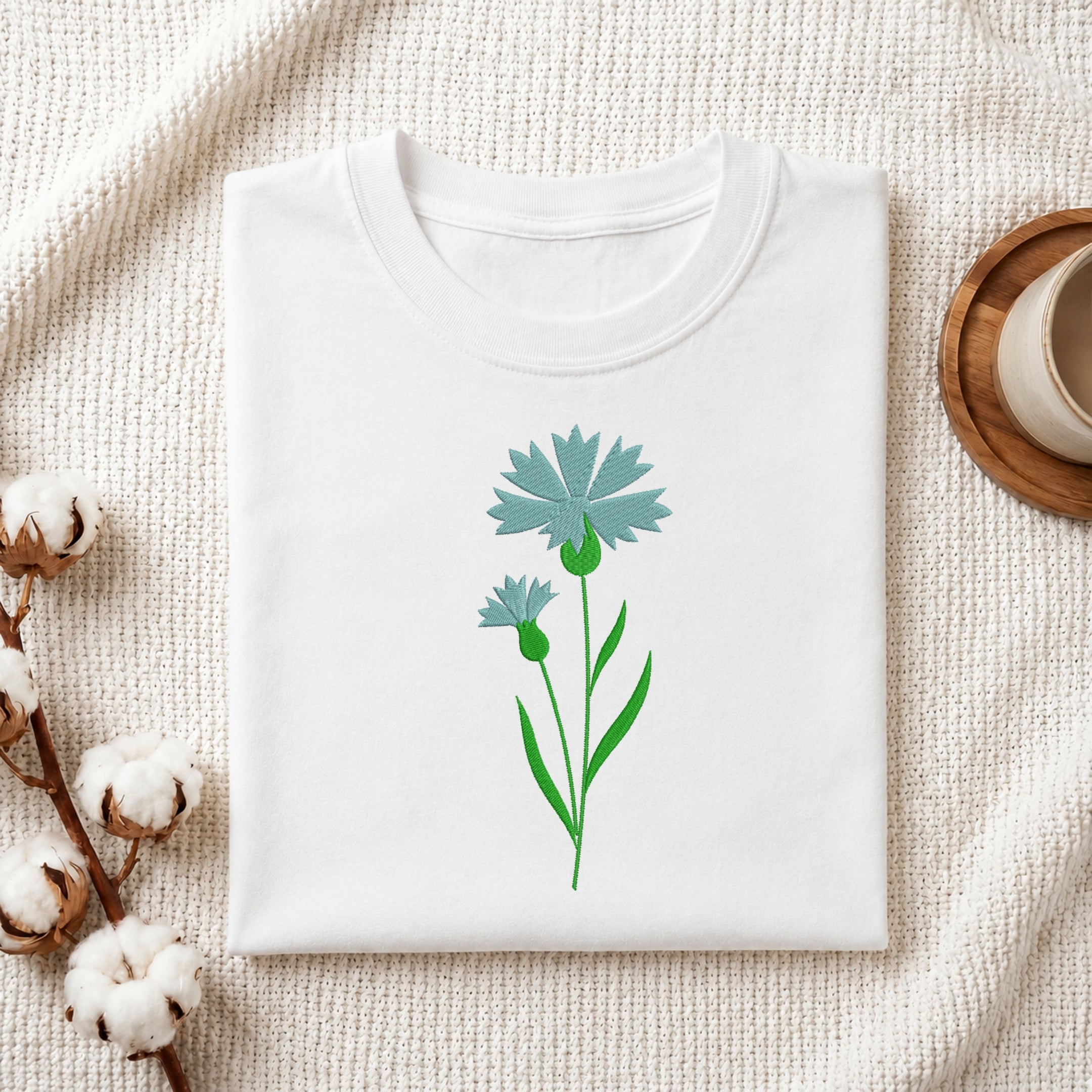 A folded white crewneck t-shirt featuring a graphic of two light blue cornflowers with green stems, styled on a textured cream knit blanket with cotton bolls and a wooden cup in the background.