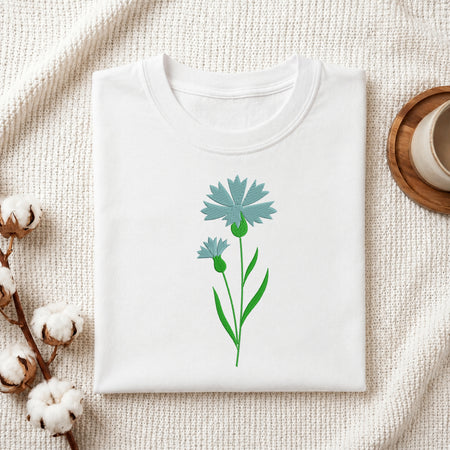 A folded white crewneck t-shirt featuring a graphic of two light blue cornflowers with green stems, styled on a textured cream knit blanket with cotton bolls and a wooden cup in the background.