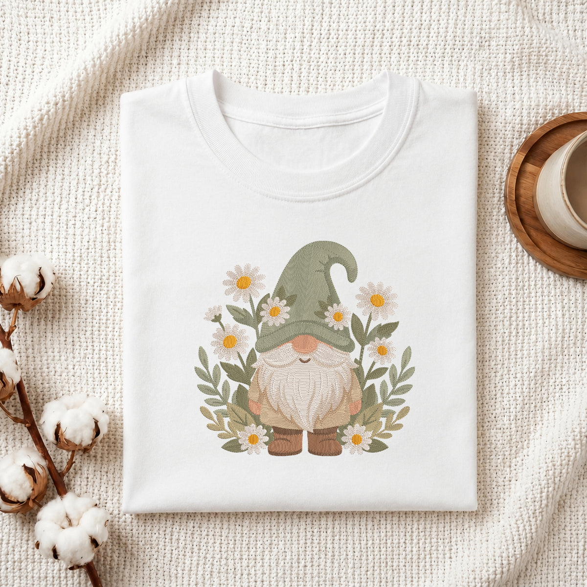 A folded white crewneck t-shirt featuring a cute daisy-and-gnome graphic, styled on a textured cream fabric with cotton bolls and a wooden mug.