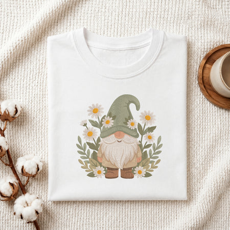 A folded white crewneck t-shirt featuring a cute daisy-and-gnome graphic, styled on a textured cream fabric with cotton bolls and a wooden mug.