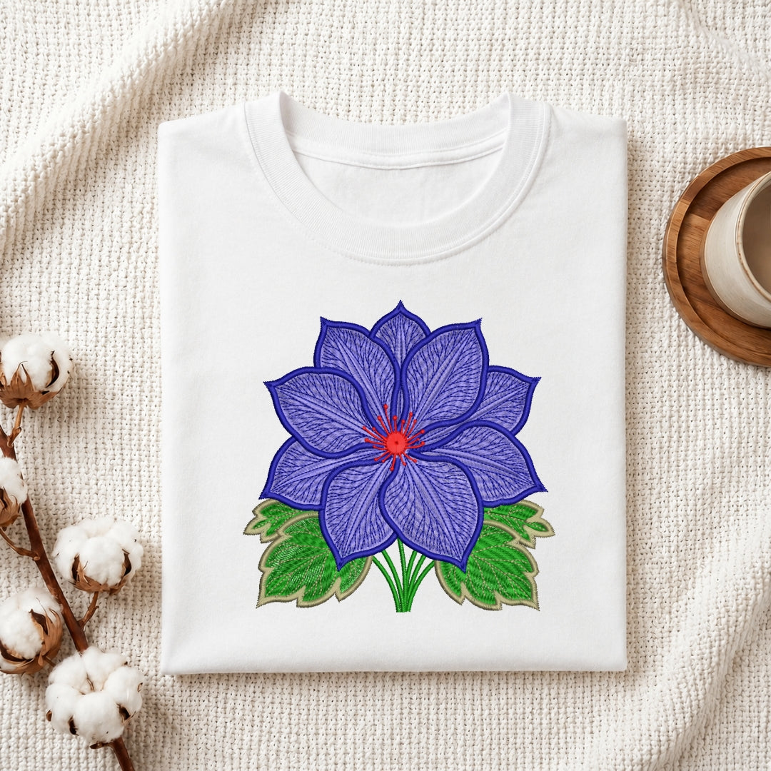 A folded white crewneck t-shirt with a colorful embroidered blue lotus flower design, displayed on a textured cream fabric background with decorative cotton bolls and a wooden cup.