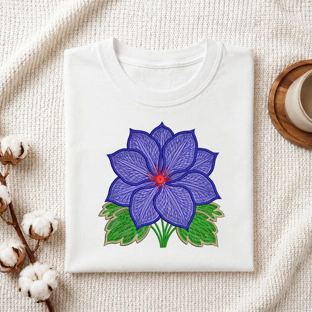 A folded white crewneck t-shirt with a colorful embroidered blue lotus flower design, displayed on a textured cream fabric background with decorative cotton bolls and a wooden cup.