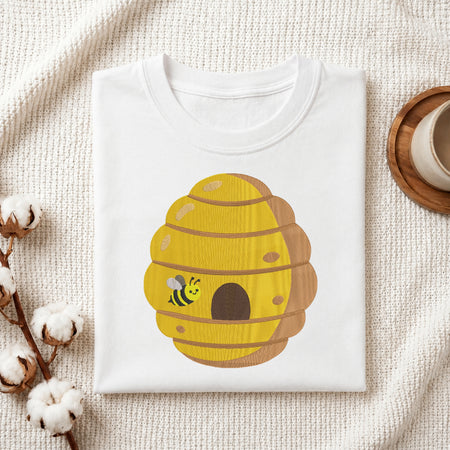 Flat-lay mockup showing a folded white sweatshirt featuring an embroidered yellow beehive with a cute bumblebee, set against a cozy white knitted blanket with cotton bolls and a wooden saucer.