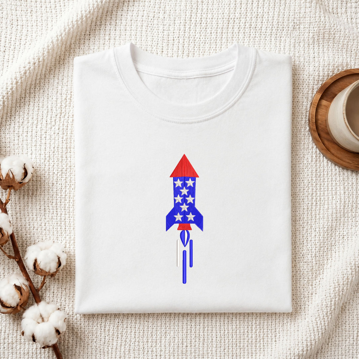 Folded white cotton t-shirt mockup featuring a patriotic space rocket star pattern machine embroidery design, styled with cotton stems and a wooden cup.