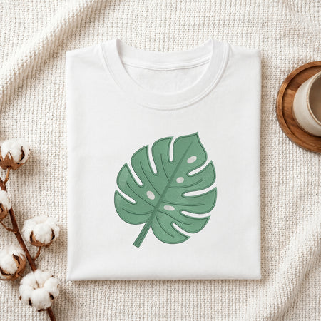 A folded white crewneck t-shirt featuring a green monstera leaf graphic on the front, styled on a textured cream knit blanket with cotton bolls and a wooden cup in the background.