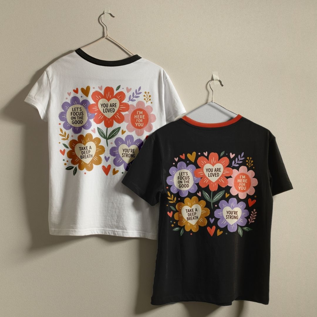 Two ringer t-shirts with colorful floral and heart graphics featuring motivational quotes, displayed on hangers against a beige wall. One shirt is white with a black collar, and the other is black with a red collar.