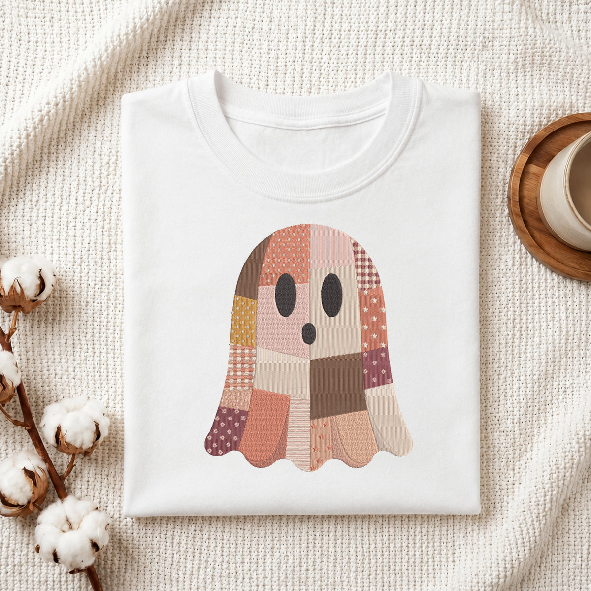 Folded white cotton t-shirt mockup featuring a patchwork Halloween ghost embroidery design, styled with cotton stems and a wooden cup.