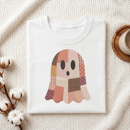 Folded white cotton t-shirt mockup featuring a patchwork Halloween ghost embroidery design, styled with cotton stems and a wooden cup.