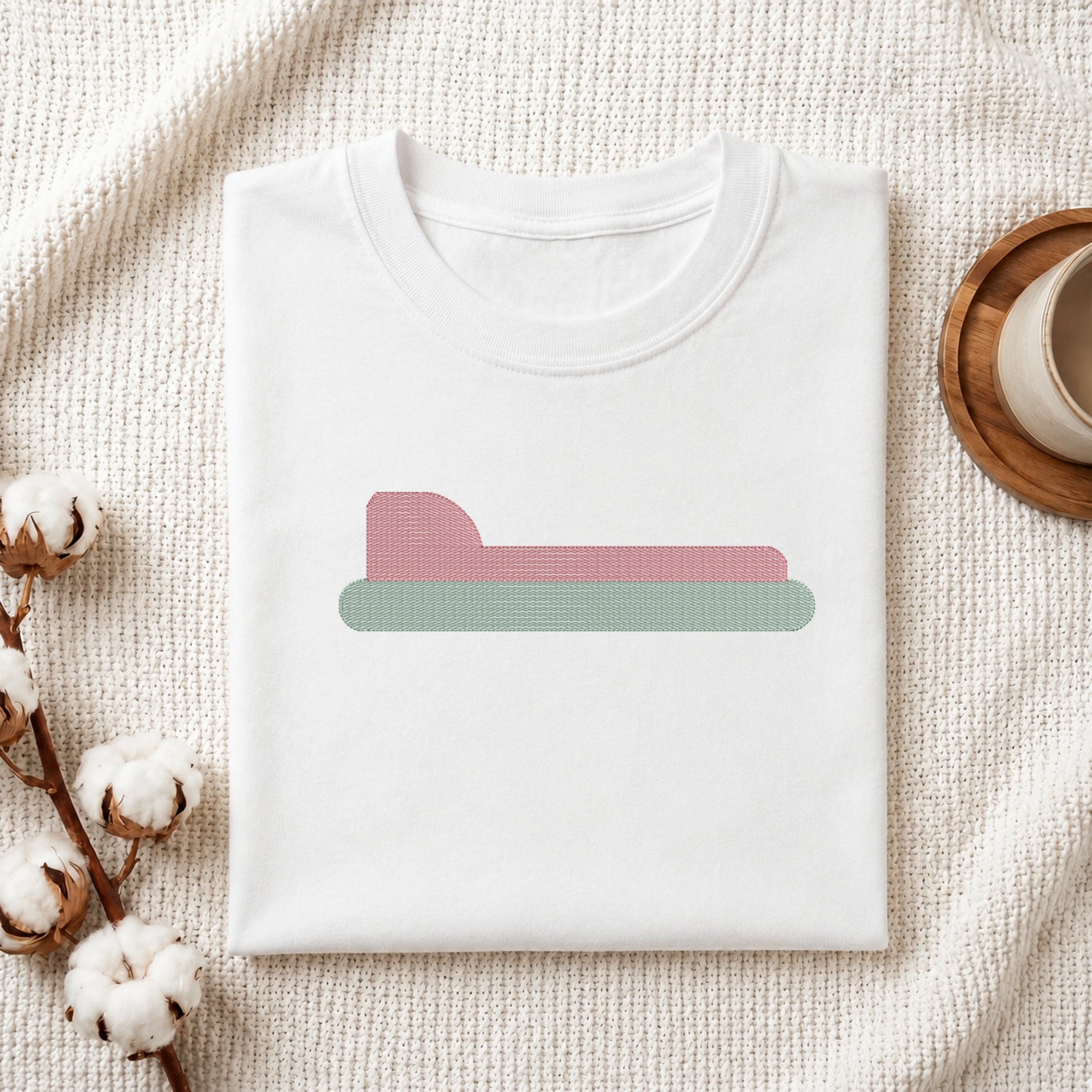 A folded white crewneck t-shirt is displayed on a textured cream knit blanket, styled with cotton bolls on the left and a wooden cup on a coaster on the right. The t-shirt features a minimalist graphic of a pink bed with a green base.