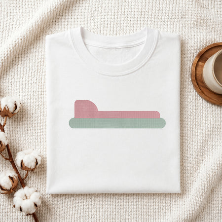 A folded white crewneck t-shirt is displayed on a textured cream knit blanket, styled with cotton bolls on the left and a wooden cup on a coaster on the right. The t-shirt features a minimalist graphic of a pink bed with a green base.