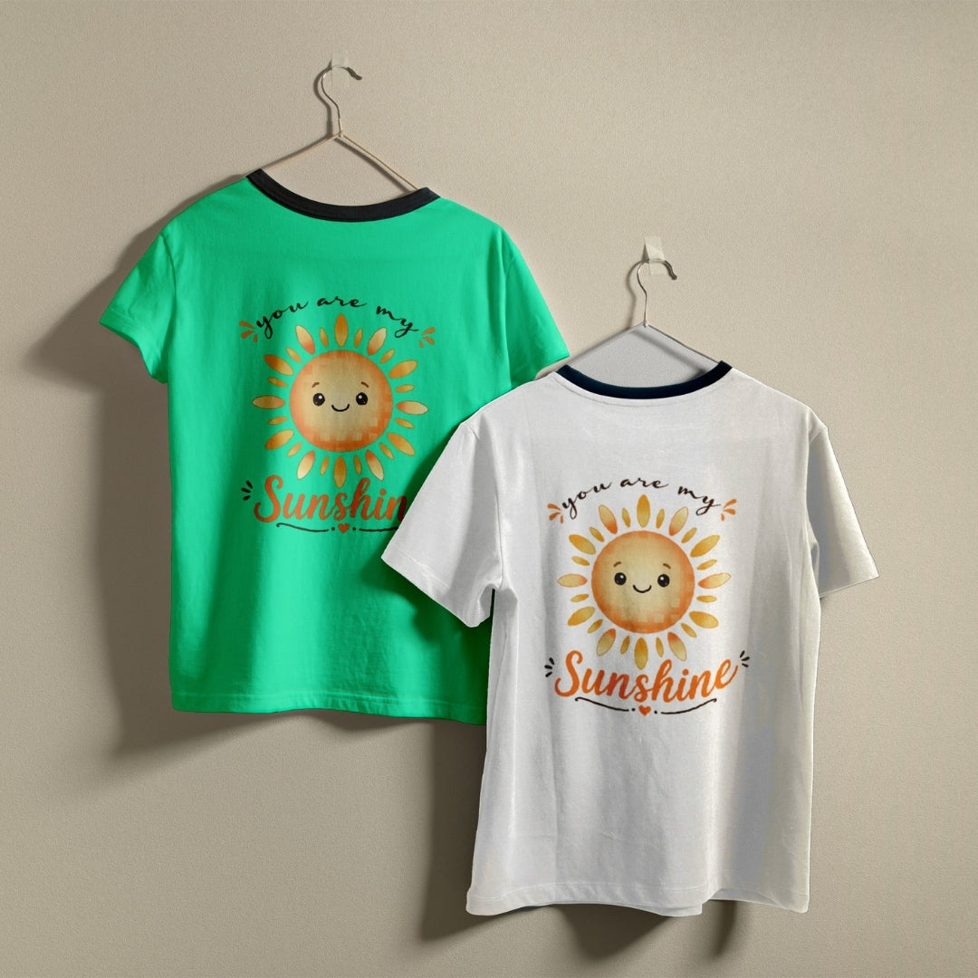 Two t-shirts hanging on hangers, one green and one white, both printed with a happy cartoon sun graphic and the text "you are my Sunshine".
