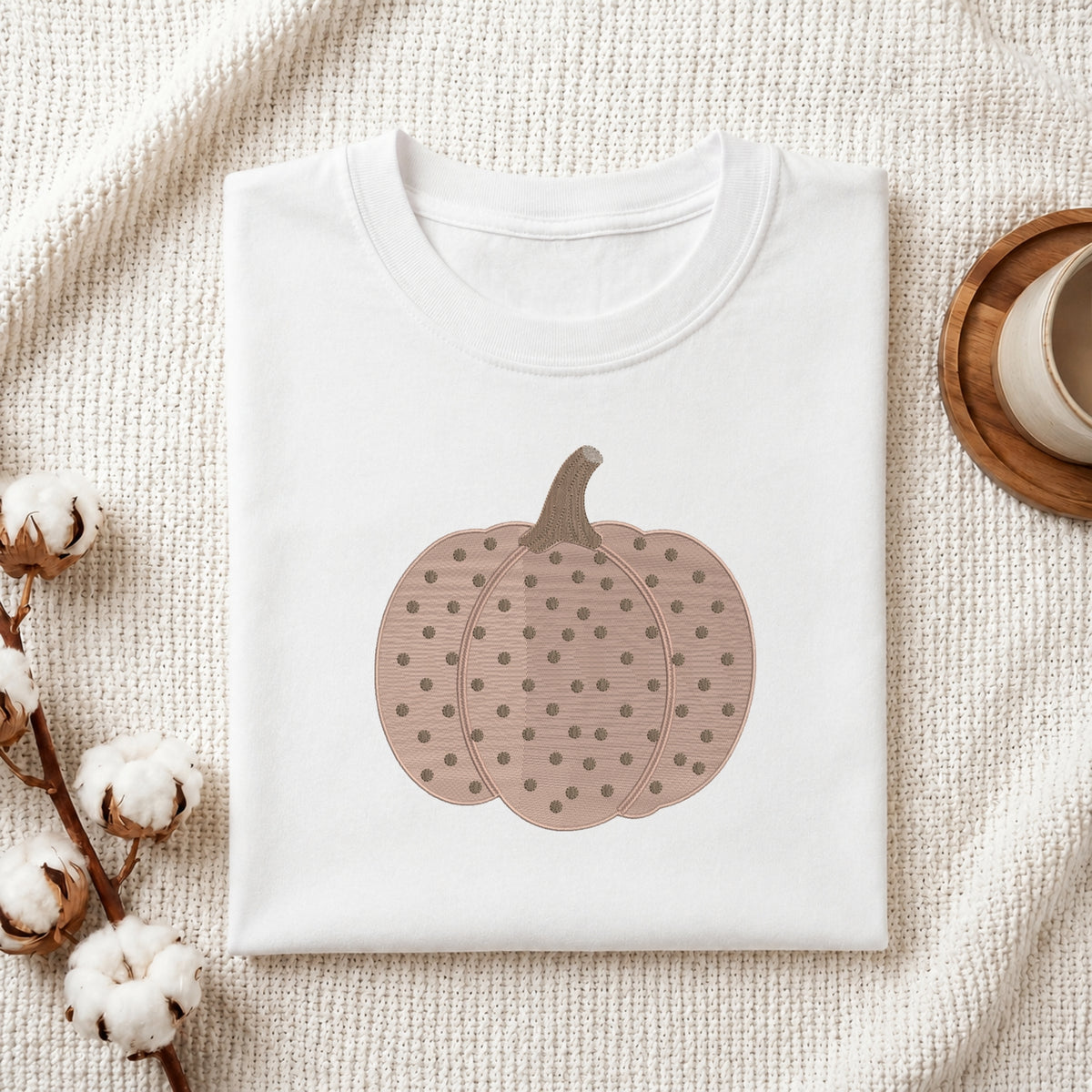 A folded white crewneck t-shirt featuring a graphic of a brown pumpkin with a polka-dot pattern, styled on a textured cream fabric with cotton stems and a wooden cup in the background.