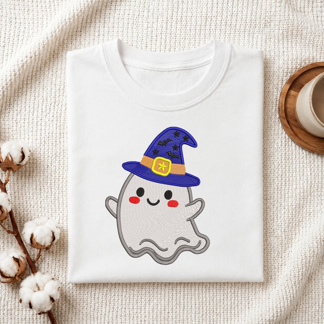 A folded white t-shirt with a cute cartoon ghost wearing a purple wizard hat, displayed on a textured cream fabric with cotton stems and a wooden cup in the background.