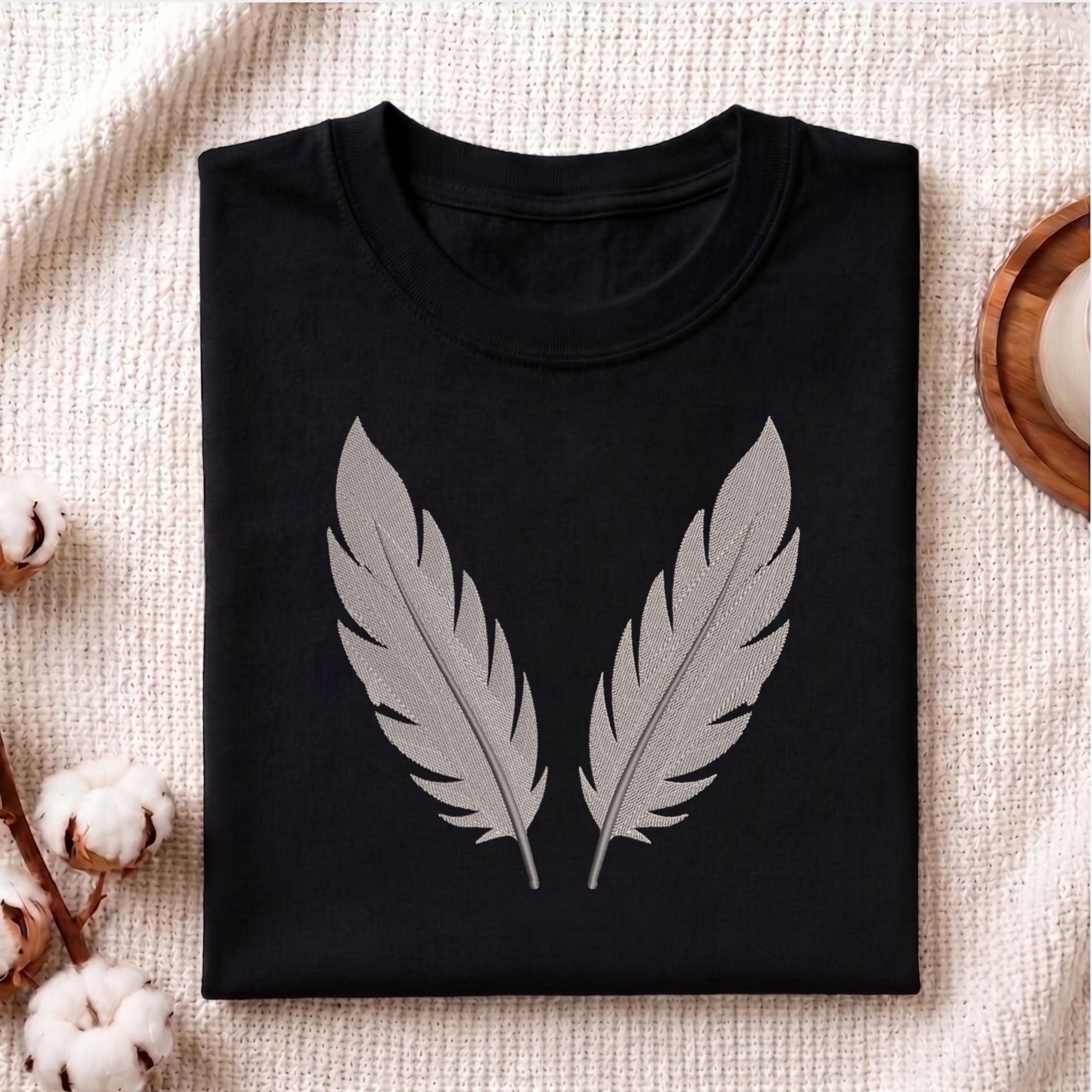 A folded black crewneck t-shirt with a large gray feather graphic on the front, displayed on a textured beige fabric background with decorative cotton bolls and a wooden tray.