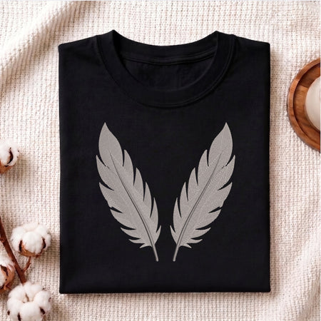 A folded black crewneck t-shirt with a large gray feather graphic on the front, displayed on a textured beige fabric background with decorative cotton bolls and a wooden tray.