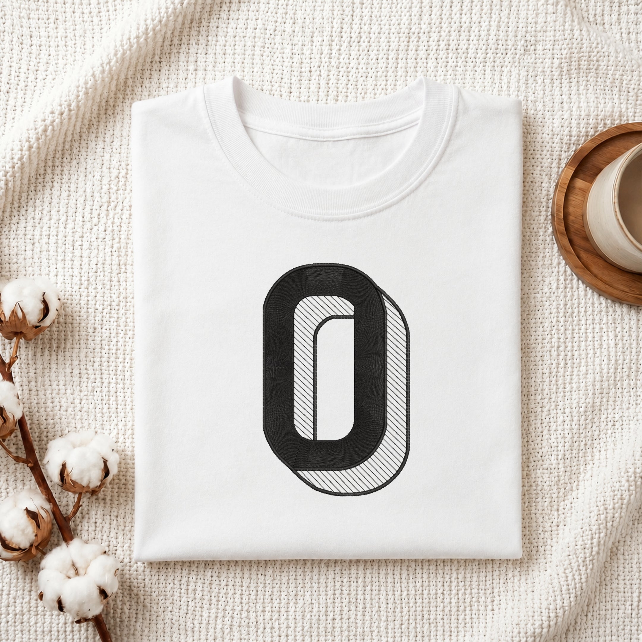 A neatly folded white crewneck t-shirt featuring a stitched 3D block letter "O" design, styled with cotton branches and a wooden coaster.
