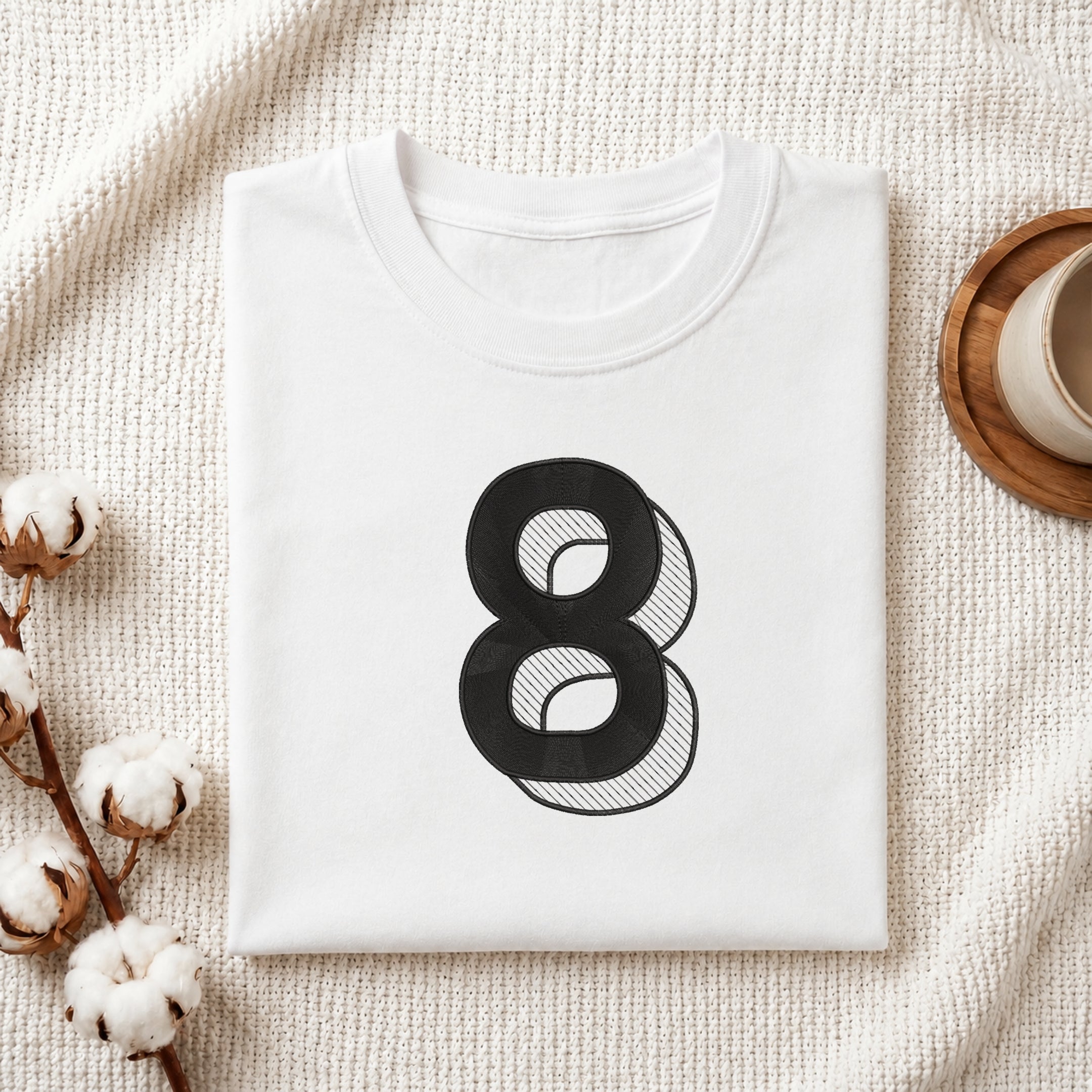 A folded white crewneck t-shirt with a large black number 8 graphic printed on the front, styled on a textured cream knit blanket with cotton stems and a wooden cup in the background.