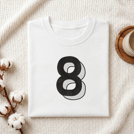 A folded white crewneck t-shirt with a large black number 8 graphic printed on the front, styled on a textured cream knit blanket with cotton stems and a wooden cup in the background.