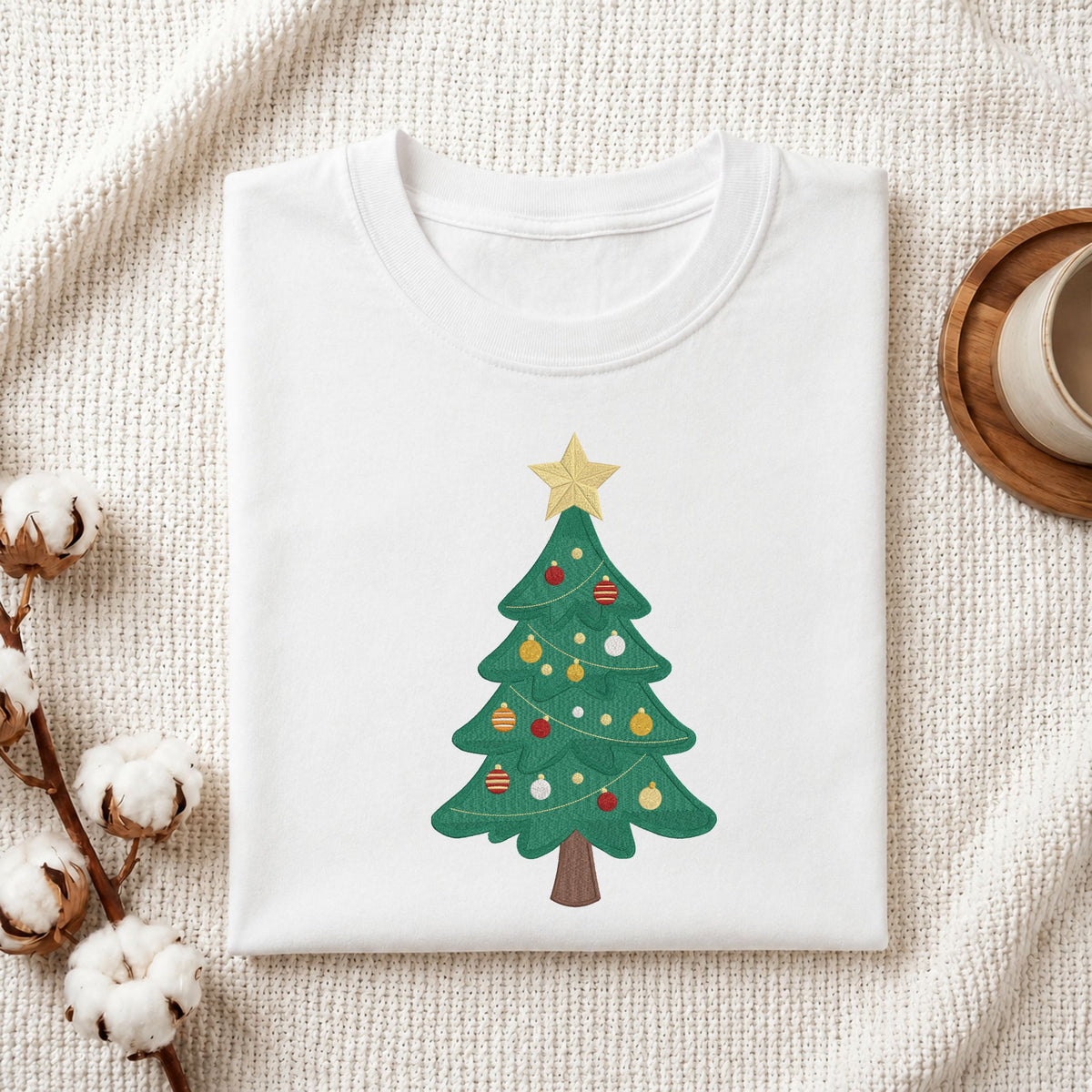 Folded white cotton t-shirt mockup featuring a classic decorated Christmas tree machine embroidery design, styled with cotton stems and a wooden cup   