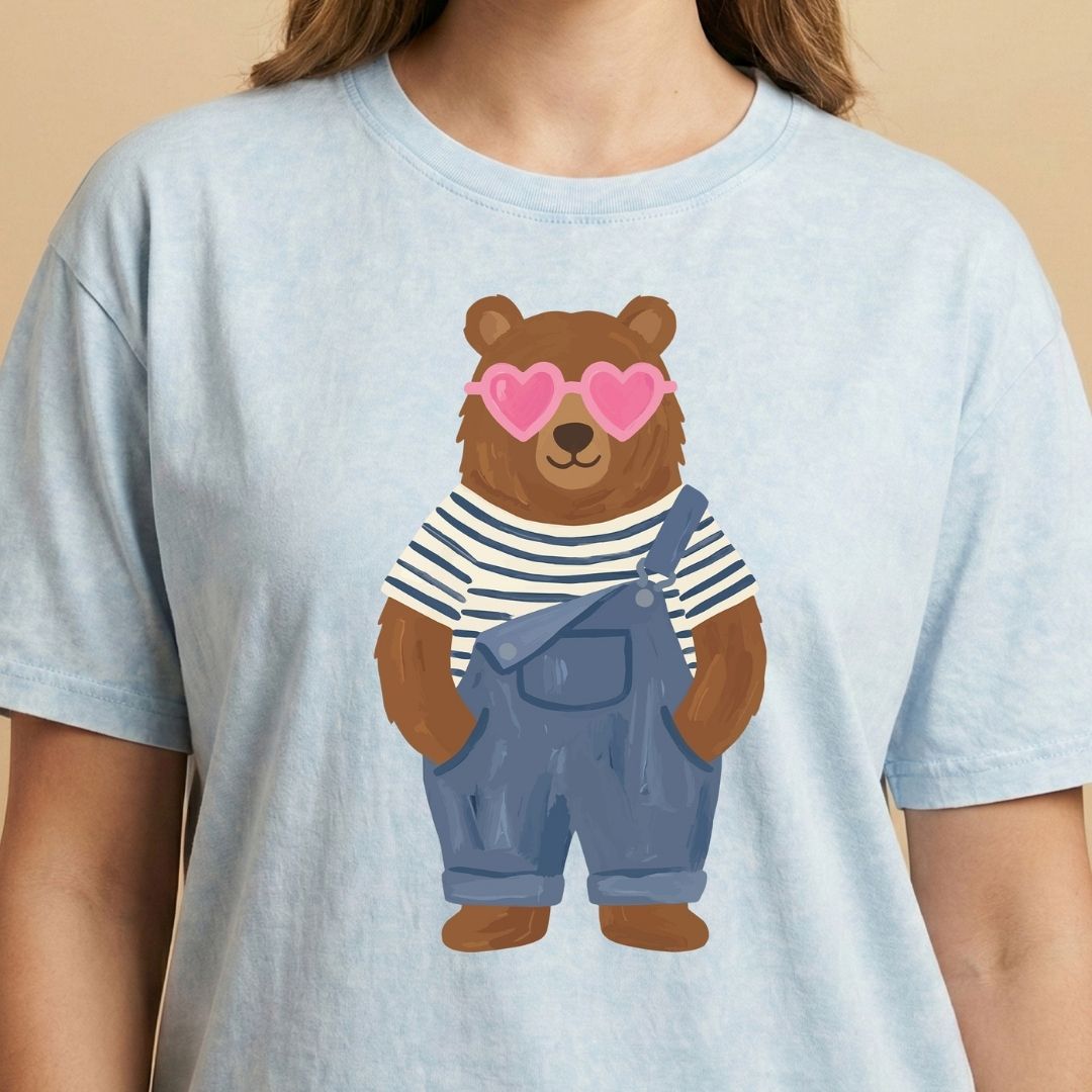 Light blue t-shirt with a bear wearing sunglasses and overalls on a beige background