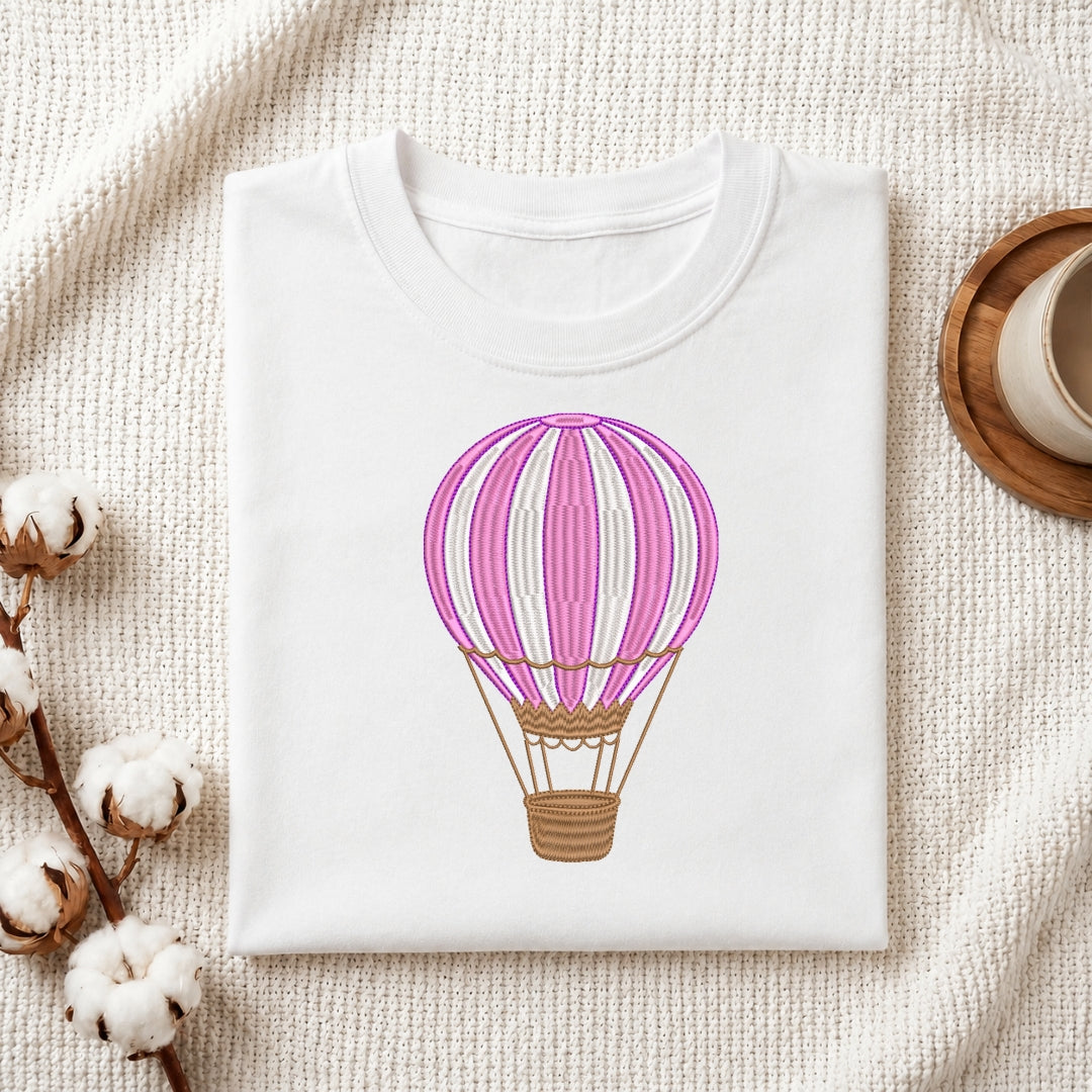 The hot air balloon embroidery design is featured on a white t-shirt, which is neatly folded on a textured white fabric.