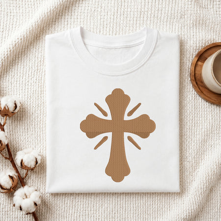A folded white T-shirt with a large brown cross graphic on the front, styled on a textured cream fabric with cotton stems and a small cup in the background.