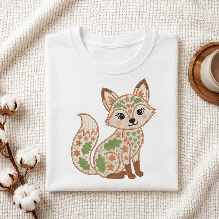 A folded white crewneck t-shirt featuring a cute embroidered-style cartoon fox with green leaves and gold stars on its fur, displayed on a textured cream knit blanket with cotton bolls and a wooden cup in the background.