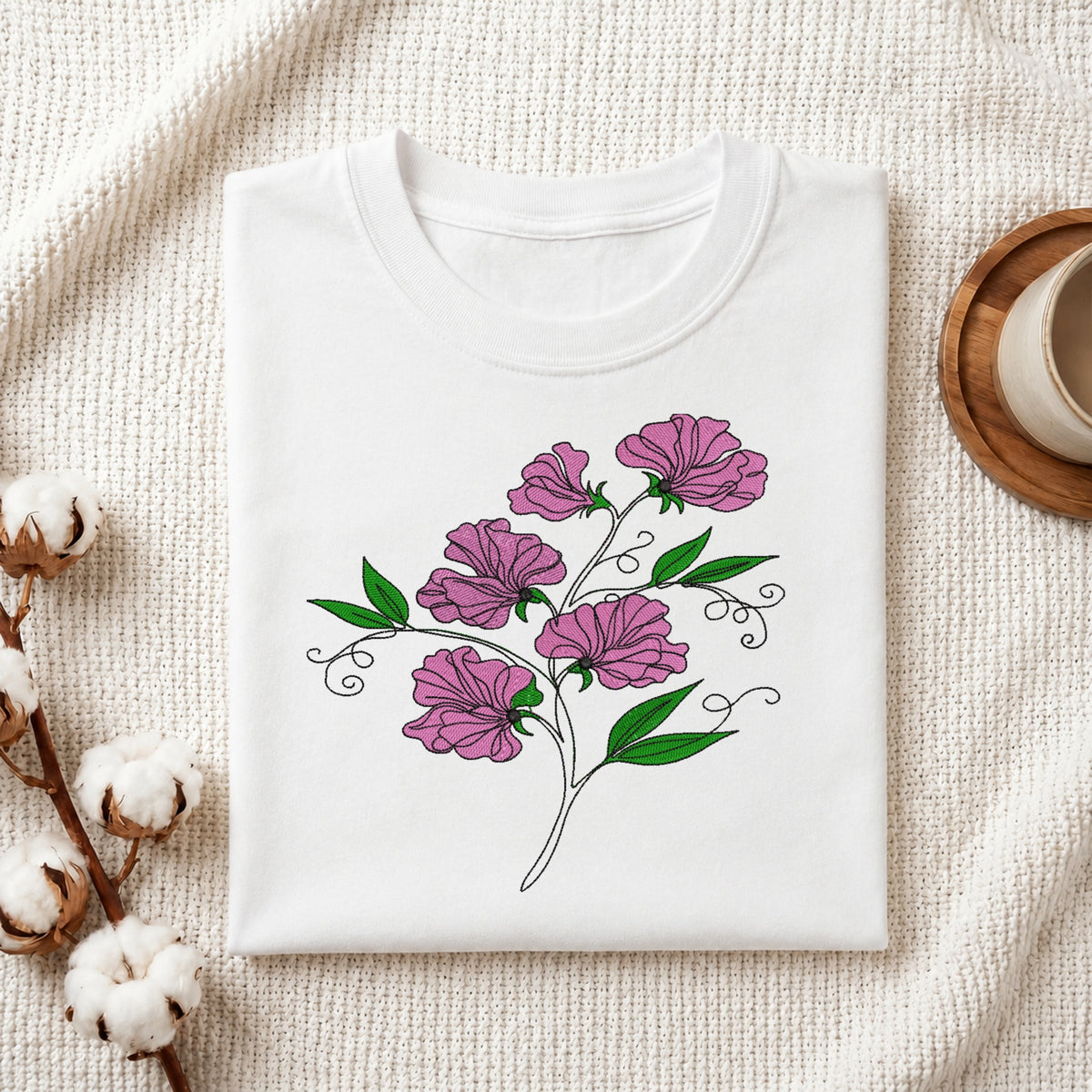 A folded white crewneck t-shirt with a graphic of purple flowers and green leaves, styled on a textured cream fabric with cotton bolls and a wooden cup.
