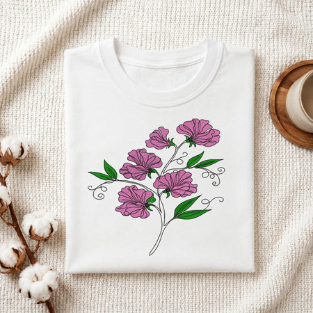 A folded white crewneck t-shirt with a graphic of purple flowers and green leaves, styled on a textured cream fabric with cotton bolls and a wooden cup.