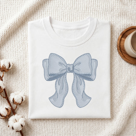 A folded white crewneck t-shirt featuring a light blue bow graphic printed on the front, styled on a textured cream knit blanket with cotton bolls and a wooden cup in the background.