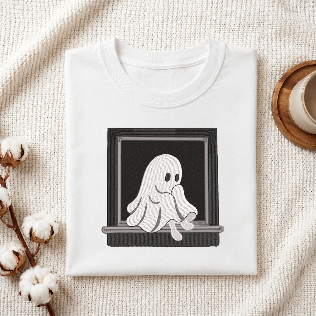 A folded white crewneck t-shirt with a graphic of a cute, wavy white ghost sitting in a black window frame, styled on a textured cream fabric with cotton bolls and a coffee cup.