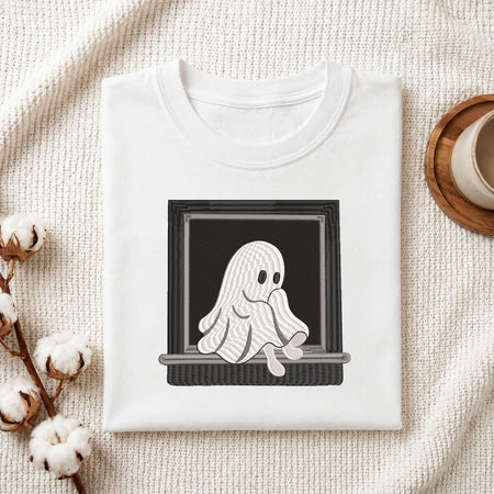 A folded white crewneck t-shirt with a graphic of a cute, wavy white ghost sitting in a black window frame, styled on a textured cream fabric with cotton bolls and a coffee cup.