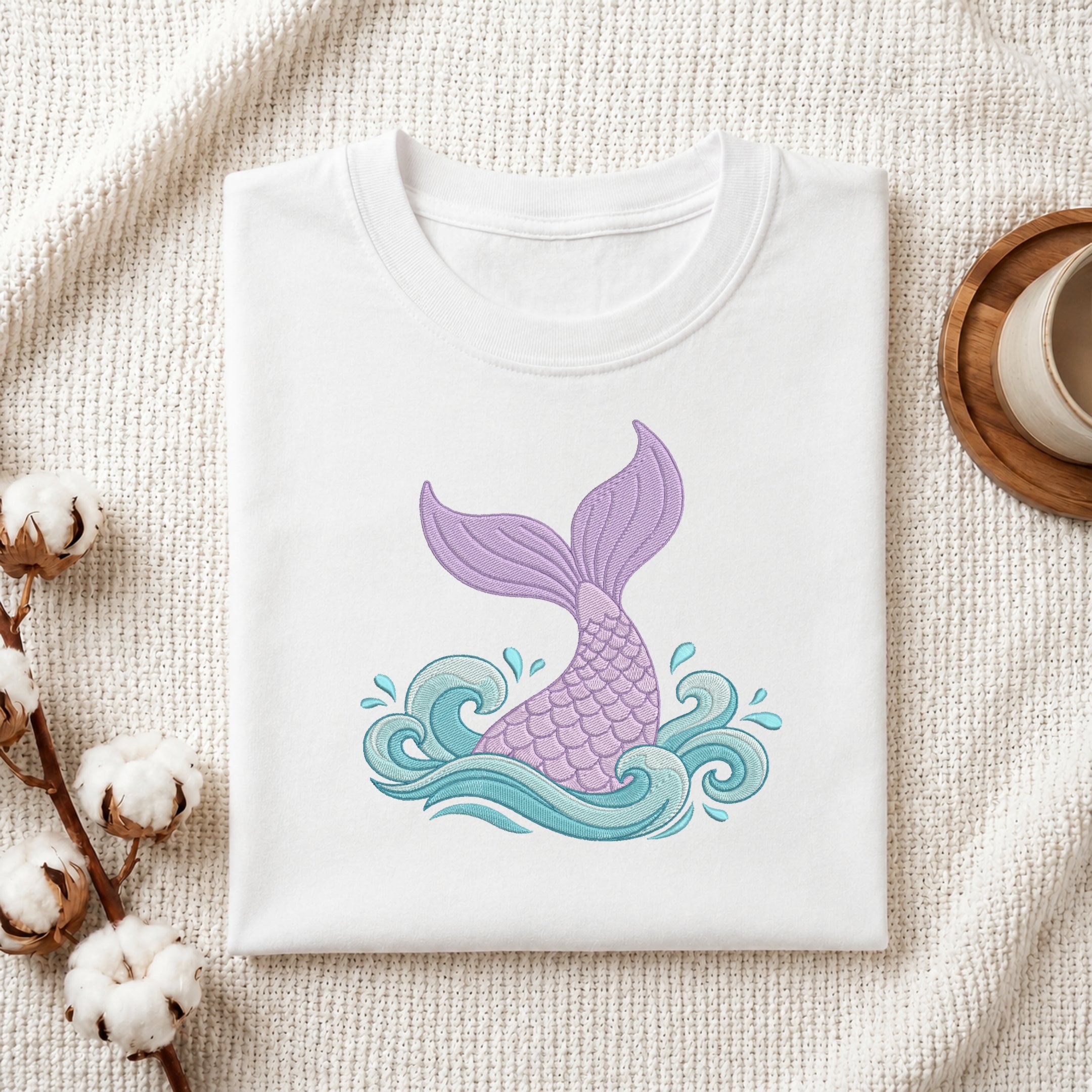 Folded white cotton t-shirt mock-up featuring a pastel purple and turquoise mermaid tail embroidery design styled with cotton branches and a ceramic mug.