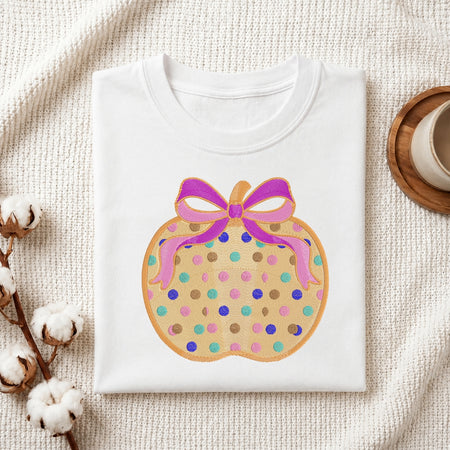 A folded white crewneck t-shirt displayed on a textured cream fabric background, decorated with cotton bolls and a wooden cup. The t-shirt features a graphic of a pumpkin cookie with a pink bow and colorful dots.