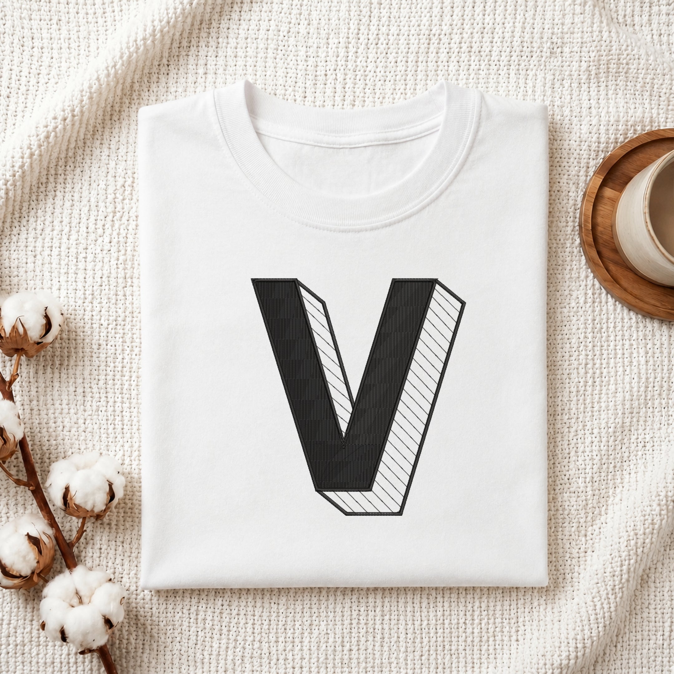 A folded white t-shirt with a large black 3D-style letter 'V' graphic on the front, styled in a flat lay on a textured cream fabric with cotton stems and a cup in the background.