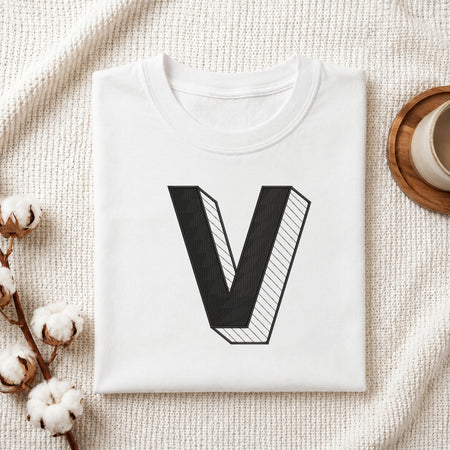 A folded white t-shirt with a large black 3D-style letter 'V' graphic on the front, styled in a flat lay on a textured cream fabric with cotton stems and a cup in the background.