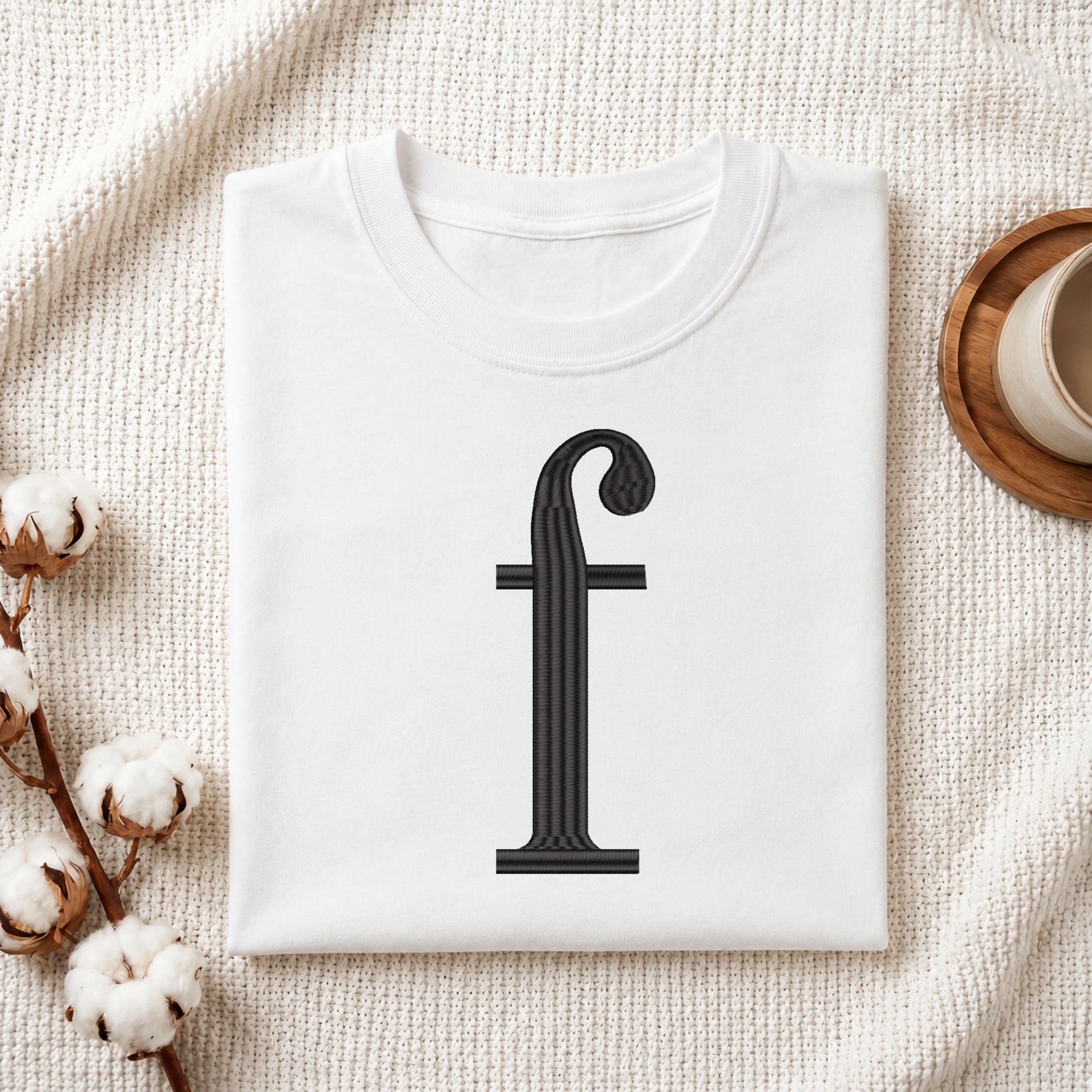 A folded white t-shirt with a black serif letter 'f' printed on the front, styled on a textured cream fabric background with cotton bolls and a cup on a wooden coaster.