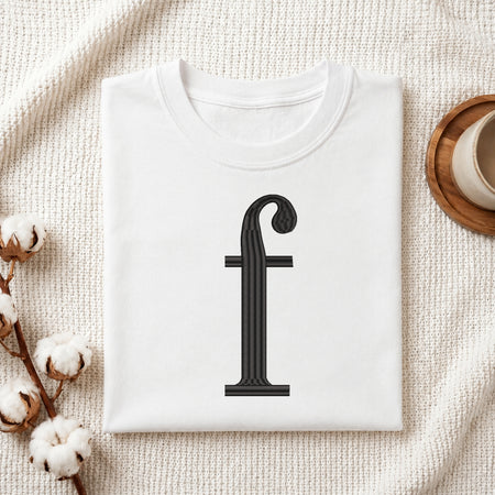 A folded white t-shirt with a black serif letter 'f' printed on the front, styled on a textured cream fabric background with cotton bolls and a cup on a wooden coaster.
