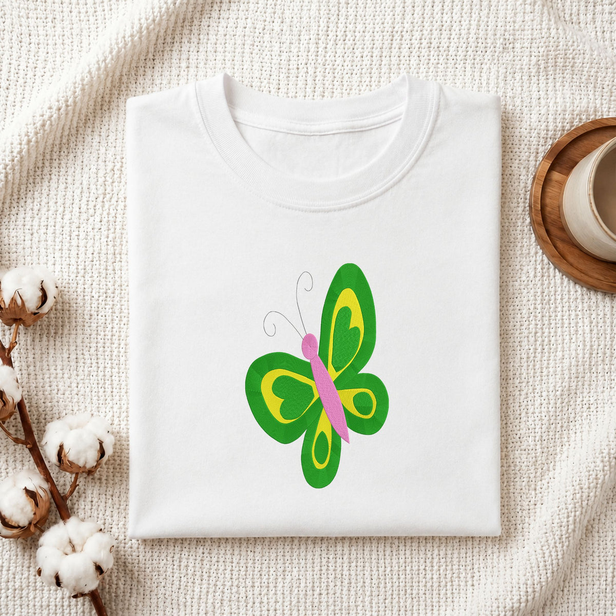 Folded white cotton t-shirt mockup featuring a butterfly machine embroidery design, styled with cotton stems and a wooden cup   