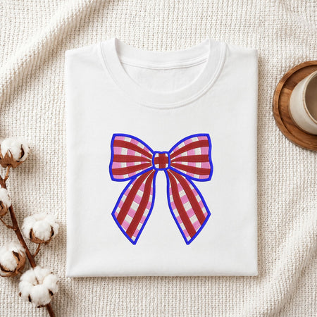 Folded white cotton t-shirt mockup featuring a red and pink plaid bow machine embroidery design, styled with cotton stems and a wooden cup