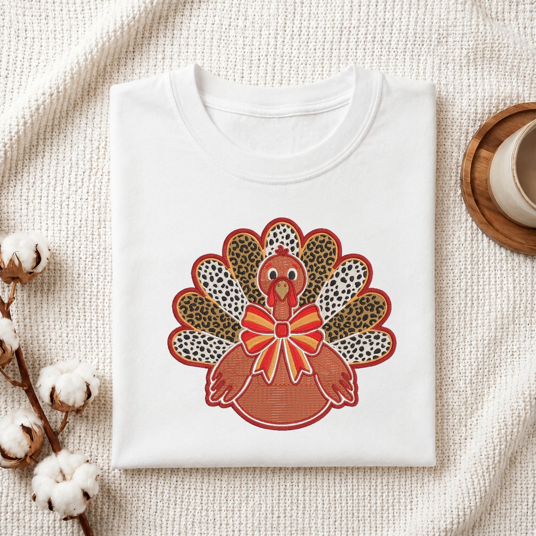 A folded white t-shirt with a colorful embroidered turkey design on the front, styled on a textured cream fabric background with cotton bolls and a wooden cup.