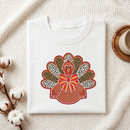 A folded white t-shirt with a colorful embroidered turkey design on the front, styled on a textured cream fabric background with cotton bolls and a wooden cup.