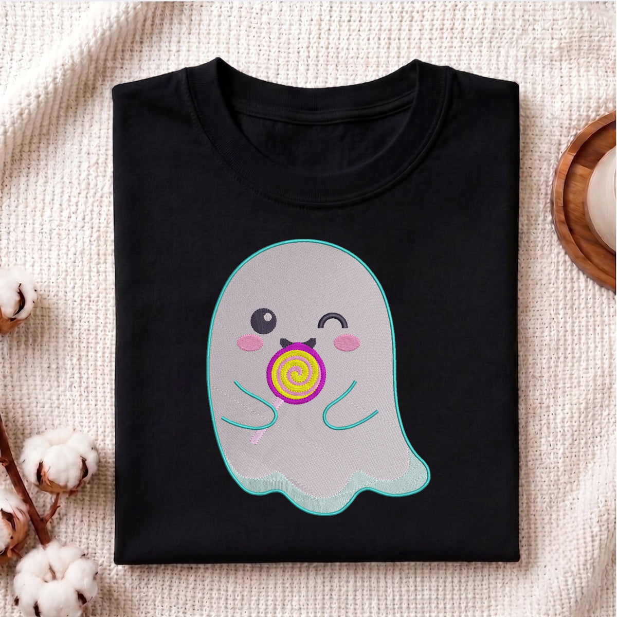 A folded black t-shirt with a cute cartoon ghost graphic holding a yellow and purple lollipop, displayed on a textured beige fabric background with cotton bolls and a wooden tray.