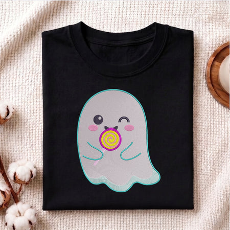 A folded black t-shirt with a cute cartoon ghost graphic holding a yellow and purple lollipop, displayed on a textured beige fabric background with cotton bolls and a wooden tray.