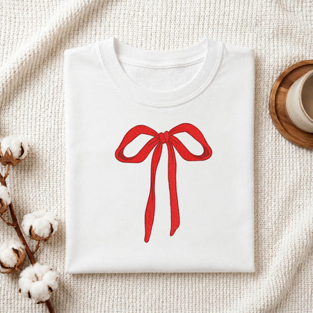 A folded white crewneck sweatshirt featuring a red ribbon bow graphic on the chest, styled on a textured cream knit blanket with cotton bolls and a wooden cup in the background.
