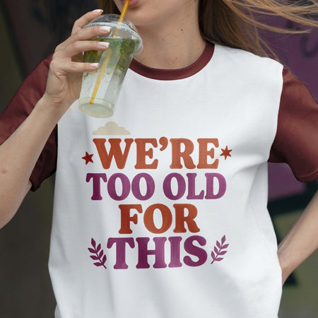 Person wearing a t-shirt with 'We're too old for this' text, holding a drink.