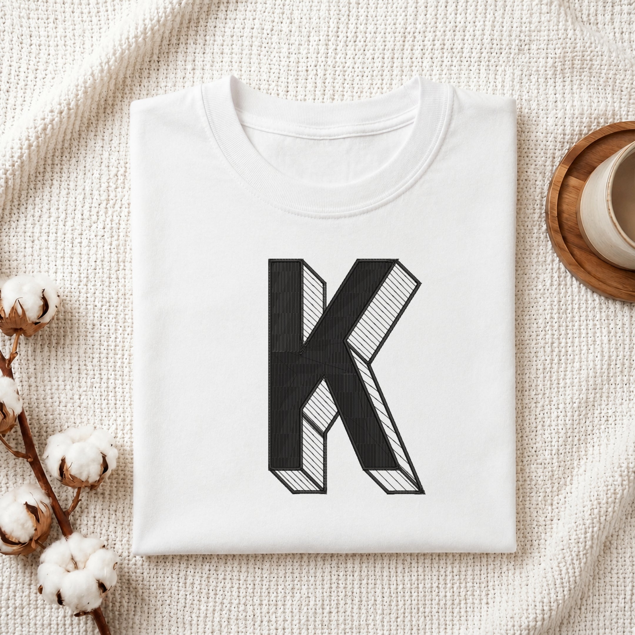 A folded white t-shirt with a large black block letter 'K' printed on the front, styled on a textured cream knit blanket with cotton bolls and a cup in the background.